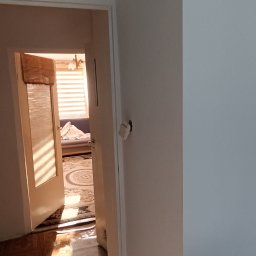 Klimek maluje - Widok z korytarza na otwarte drzwi do pokoju z roletami bambusowymi i dywanem z motywem koła, ściany w jasnych odcieniach bieli.