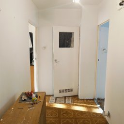 Klimek maluje - Korytarz w trakcie odświeżania w Kołobrzegu: białe ściany, brązowa podłoga w kwadraty, drzwi z szybką, widoczne narzędzia na szafce i częściowo zakryty dywanik pod drzwiami.