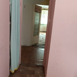 Klimek maluje - Widok z korytarza na pokój z widoczną podłogą w brązowych odcieniach, różowe ściany i drzwi z zasłoną.