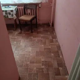 Pokój z brązową podłogą z płytek imitujących kamień, różowe ściany, okno z białymi firankami i drewniane krzesło. Widoczne rury grzewcze przy ścianie.