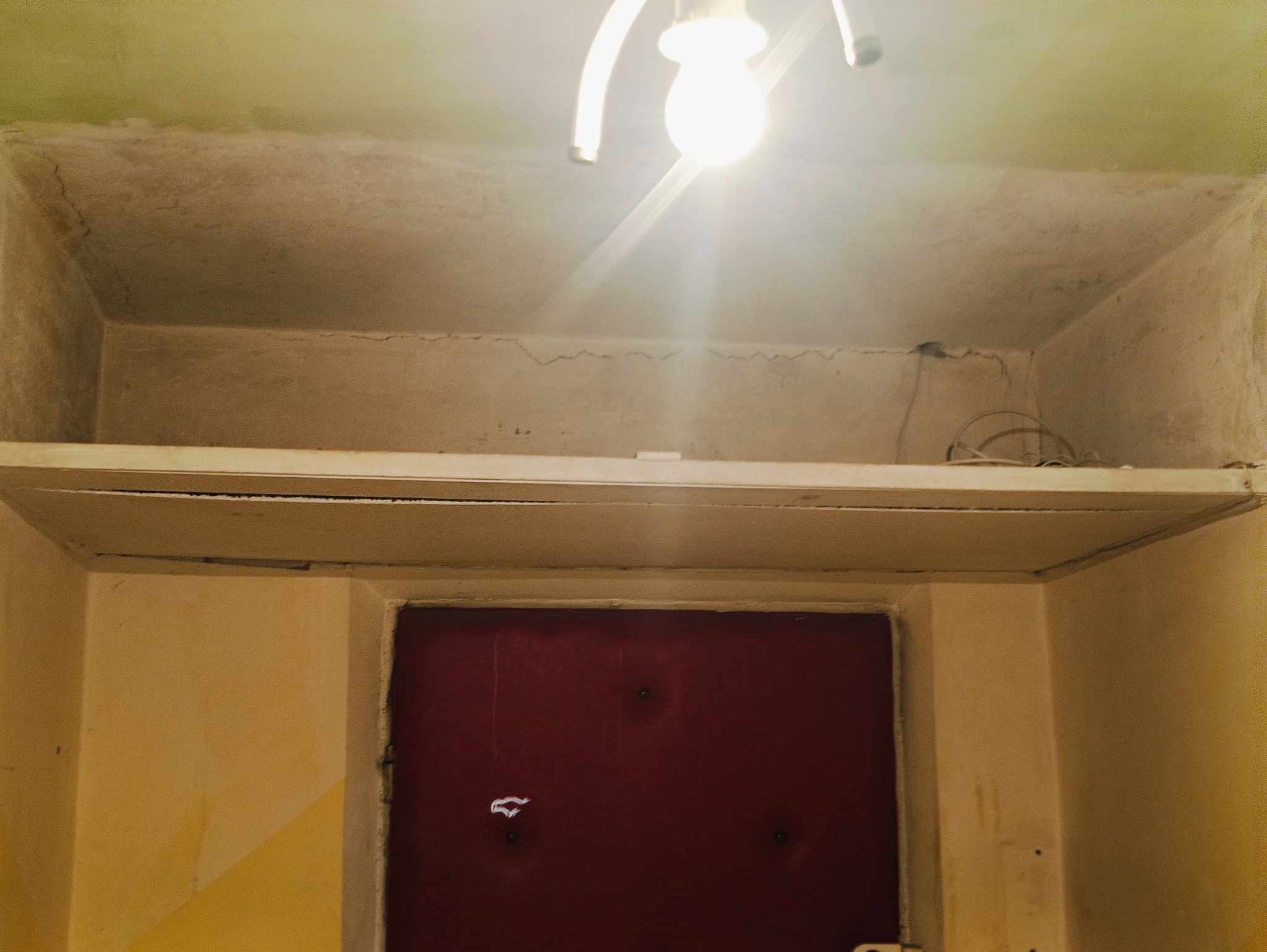 Korytarz przed pracami: surowy strop z widocznymi pęknięciami, półka pod sufitem z plątaniną kabli, ściany z resztkami farby i bordowe drzwi.