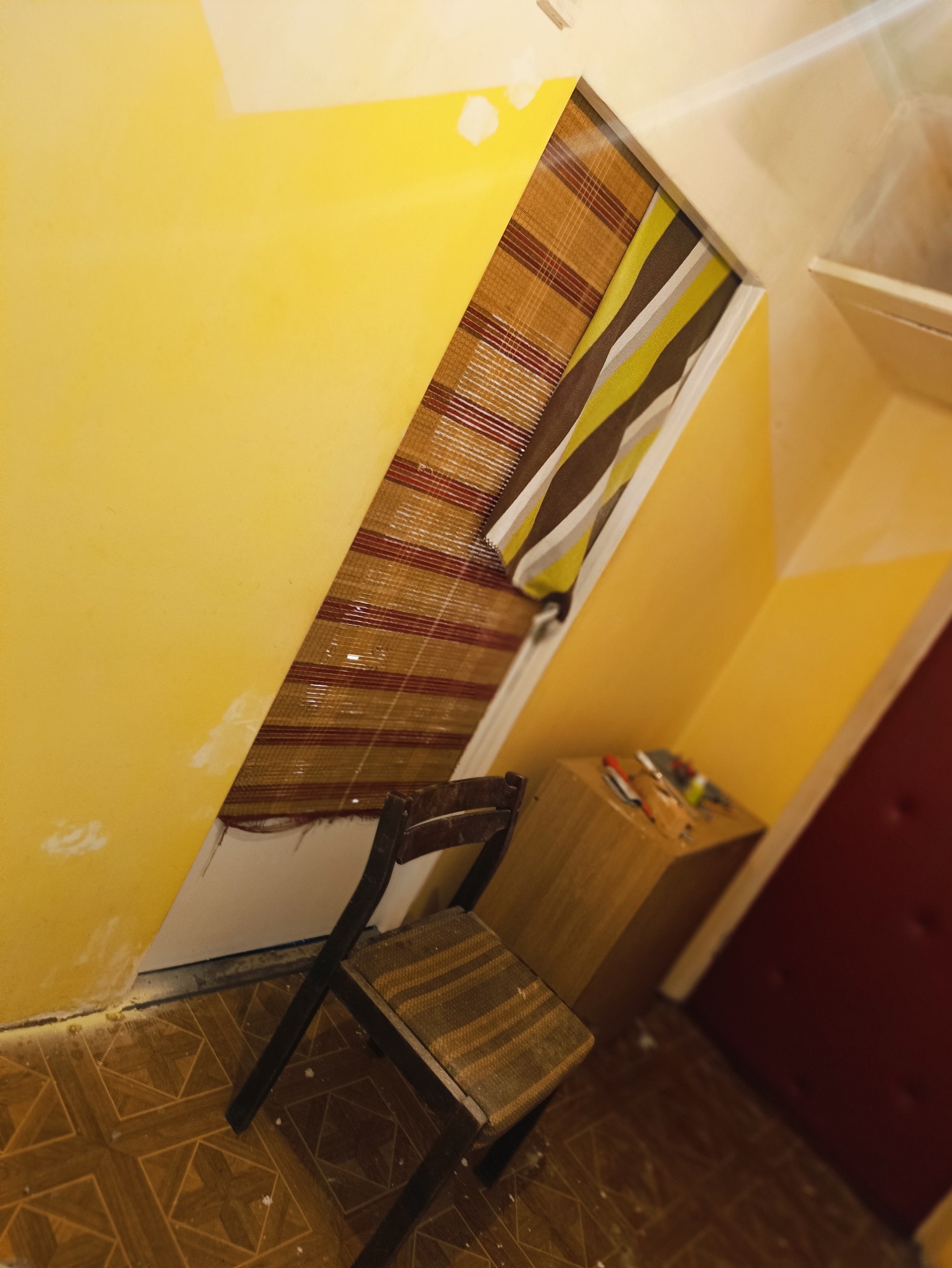 Żółta ściana w trakcie przygotowania do malowania, widoczne ślady szpachli, krzesło z brązowym siedziskiem, narzędzia na szafce i roleta w oknie, podłoga z brązowych płytek.