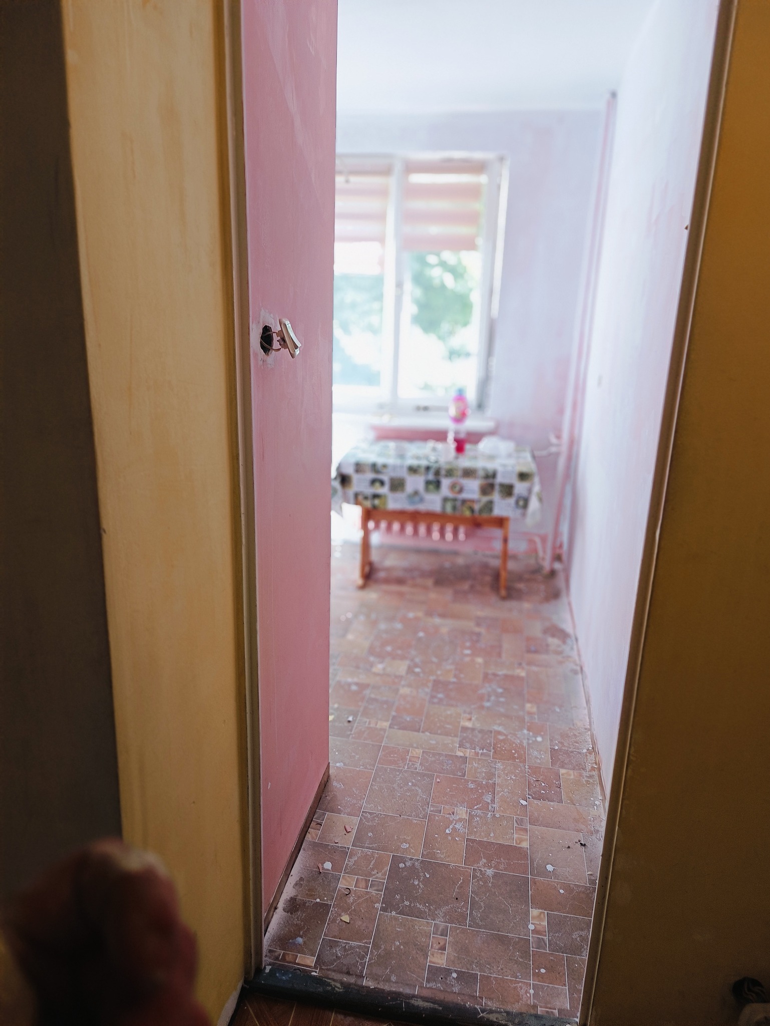 Wnętrze mieszkania w trakcie odświeżania, widok przez otwarte drzwi na pokój z pomalowanymi na różowo ścianami i brązową podłogą w płytkach, z widocznym stołem nakrytym wzorzystym obrusem przy oknie.