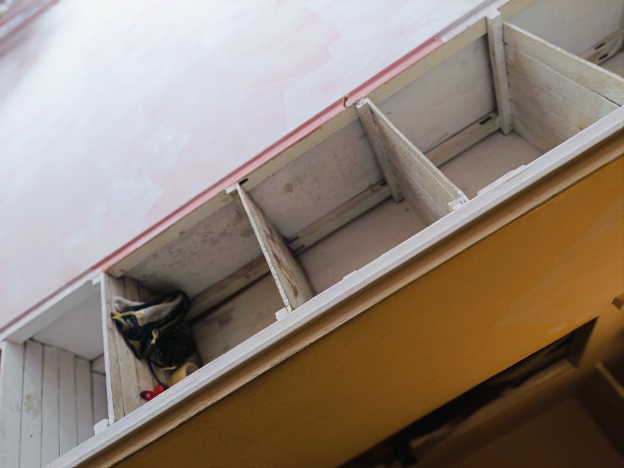 Widok z dołu na drewniany regał z półkami, świeżo pomalowany na biało, z widocznymi śladami farby na ścianie w tle, w jednej z przegródek leżą robocze rękawice.