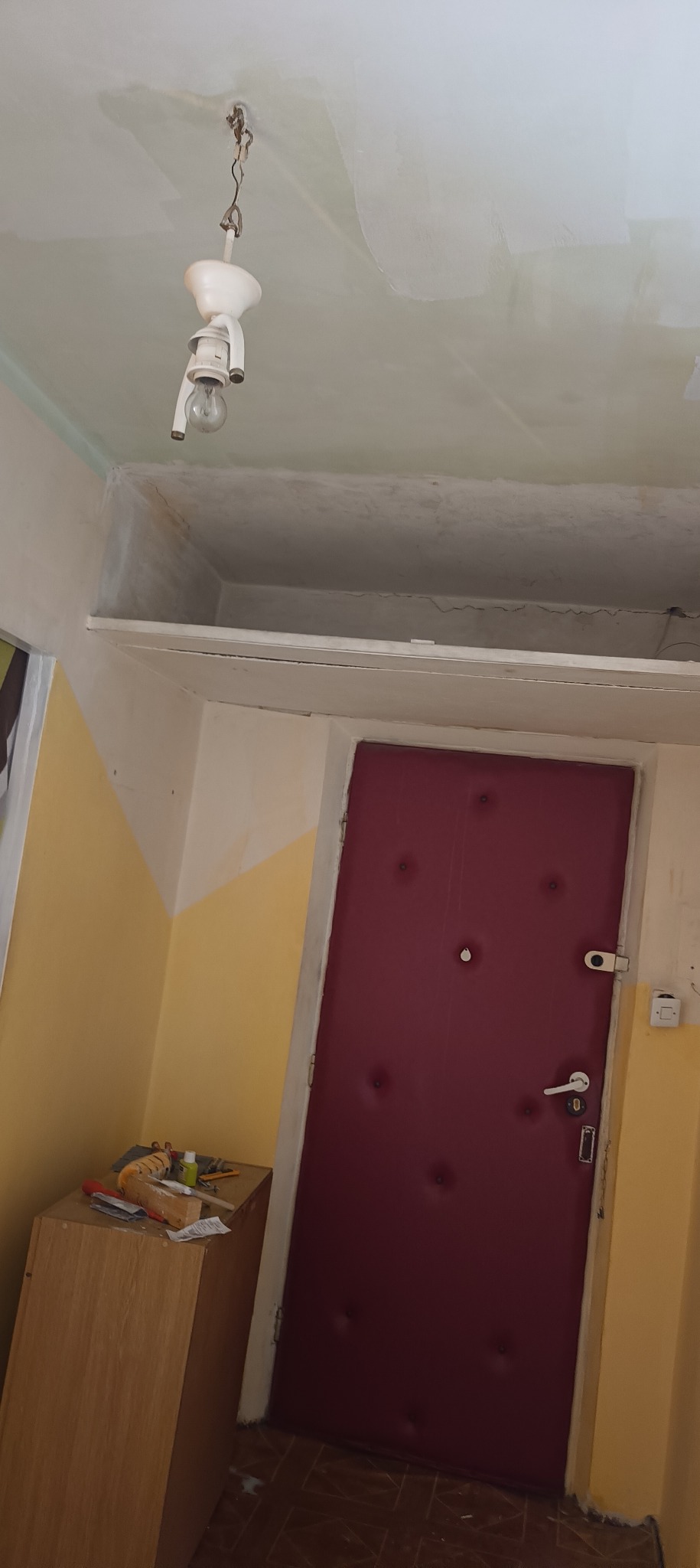 Pokój w trakcie odświeżania: sufit z widocznymi śladami gładzi, wisząca żarówka na kablu, częściowo pomalowane ściany w kolorach żółtym i białym, drzwi wejściowe w odcieniu bordo.
