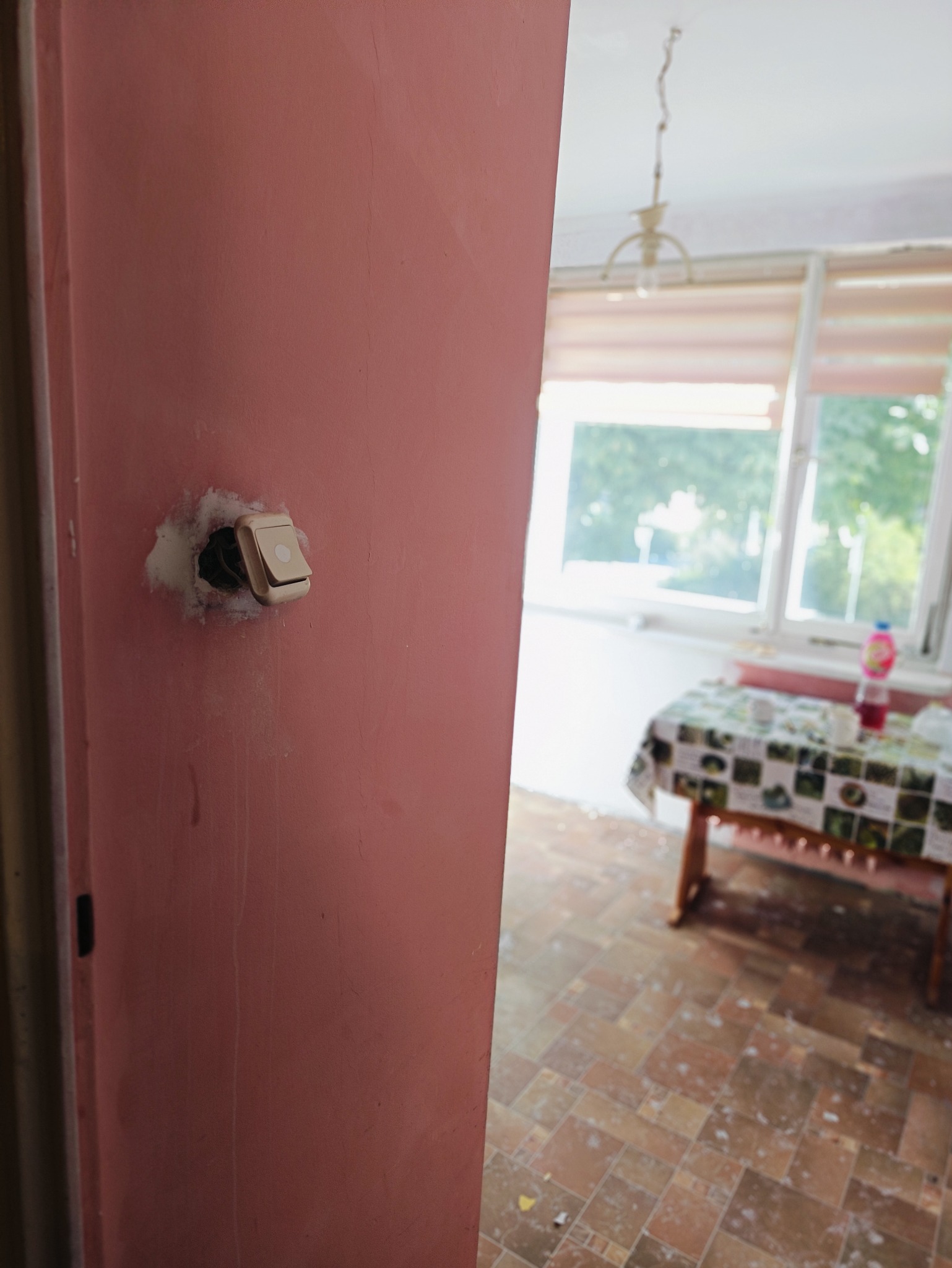 Różowa ściana z wystającym, niedbale zamontowanym włącznikiem światła, widok na pokój z oknem, stołem nakrytym ceratą i brudną podłogą.