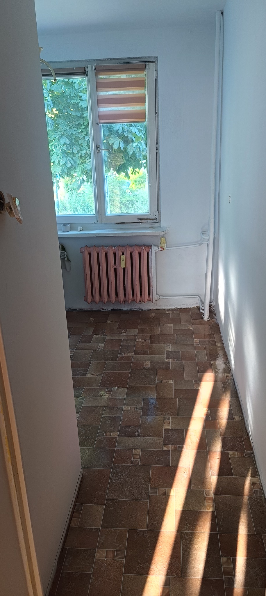 Pokój w trakcie remontu z brązową podłogą w kwadraty, różowy grzejnik pod oknem z roletami w kolorze beżowo-brązowym, białe ściany i rury oraz silne promienie słoneczne na podłodze.