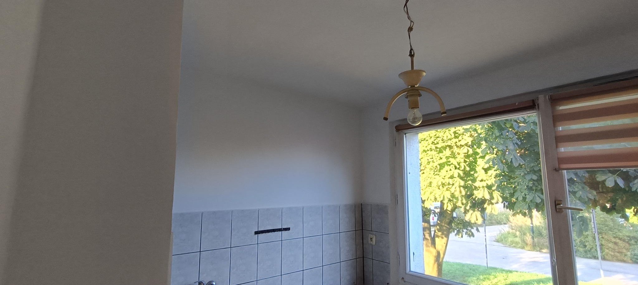 Wnętrze kuchni w trakcie odświeżania: ściany w jasnym odcieniu, fragment ściany z jasnymi kwadratowymi płytkami, widoczne ślady po demontażu elementów wyposażenia, wisząca lampa z żarówką i okno...