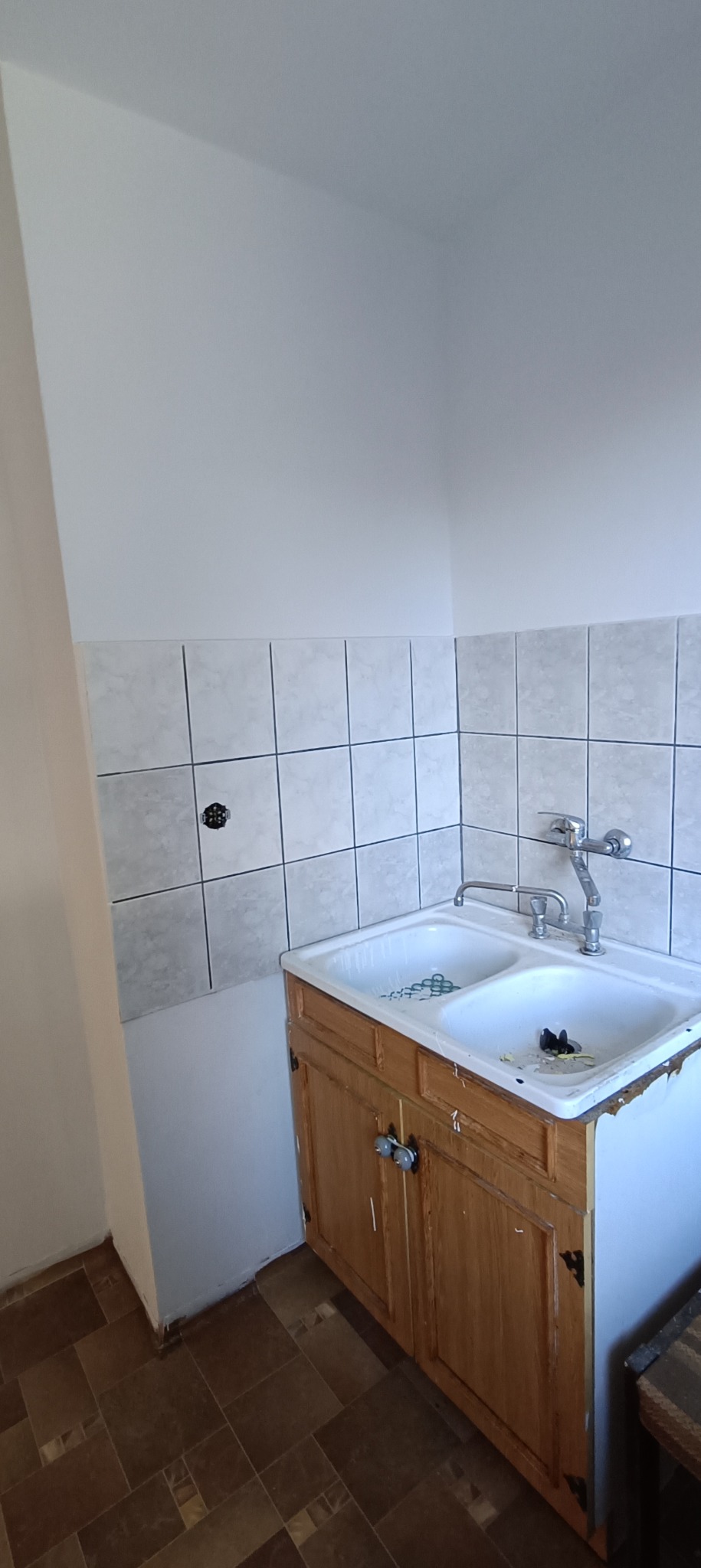 Dwukomorowy zlew w drewnianej szafce z ceramiczną okładziną ścienną w trakcie prac wykończeniowych, widoczne niedoskonałości i brakujące elementy wykończenia.