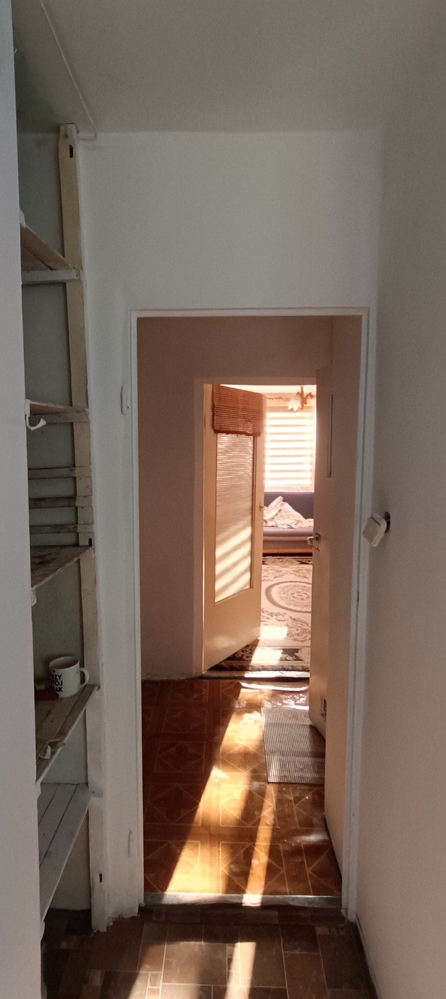 Widok z korytarza na otwarte drzwi do pokoju z dywanem i sofą, promienie słoneczne padają na podłogę, po lewej stronie widoczny regał z kubkiem.