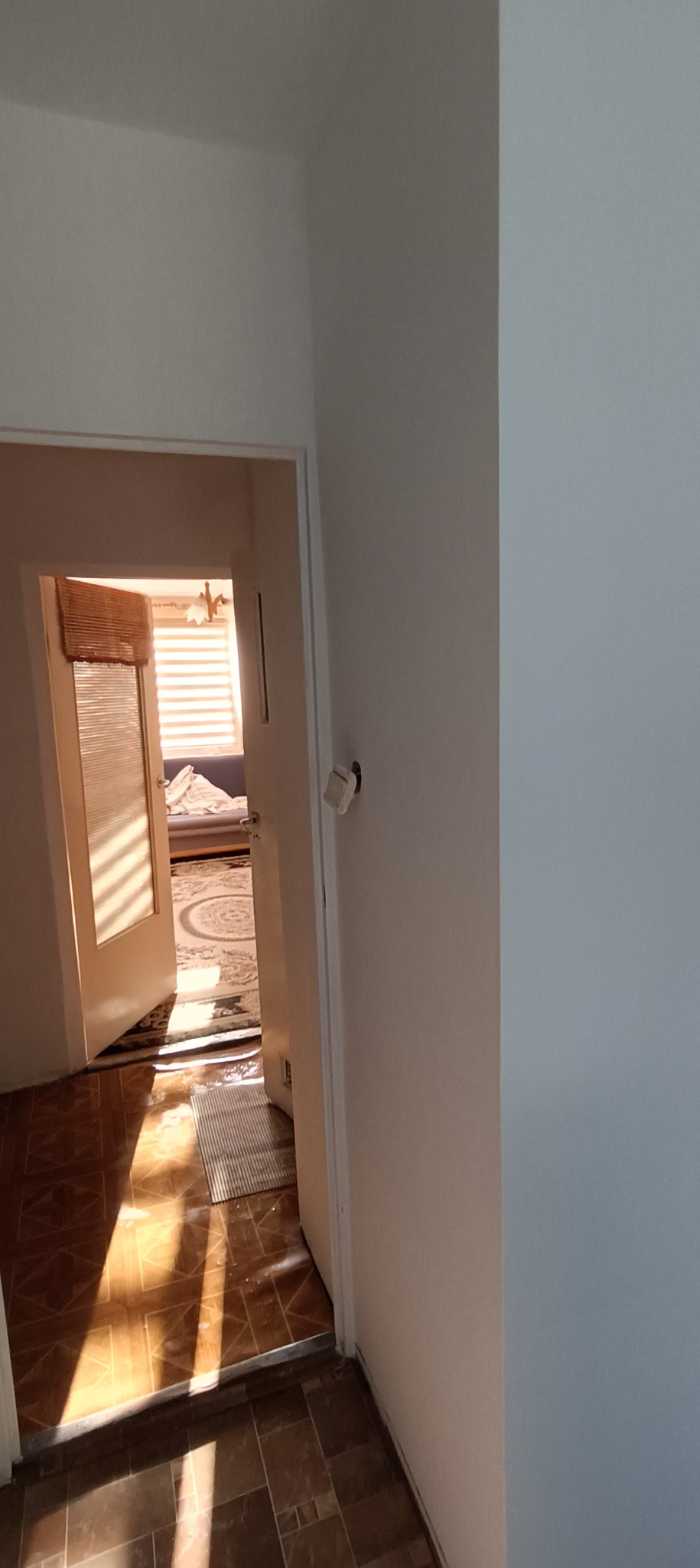 Widok z korytarza na otwarte drzwi do pokoju z roletami bambusowymi i dywanem z motywem koła, ściany w jasnych odcieniach bieli.