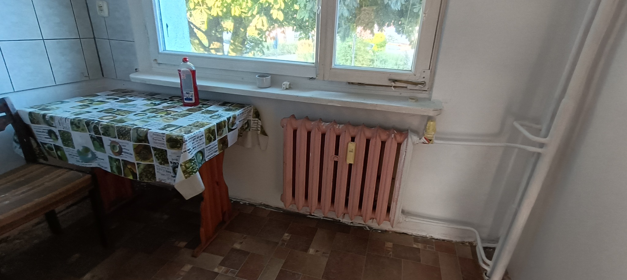 Pokój z pomalowanym na różowo grzejnikiem żeberkowym pod oknem, przy którym stoi stół nakryty ceratą w motywy kawowe, widoczny fragment brudnej podłogi i rur centralnego ogrzewania.