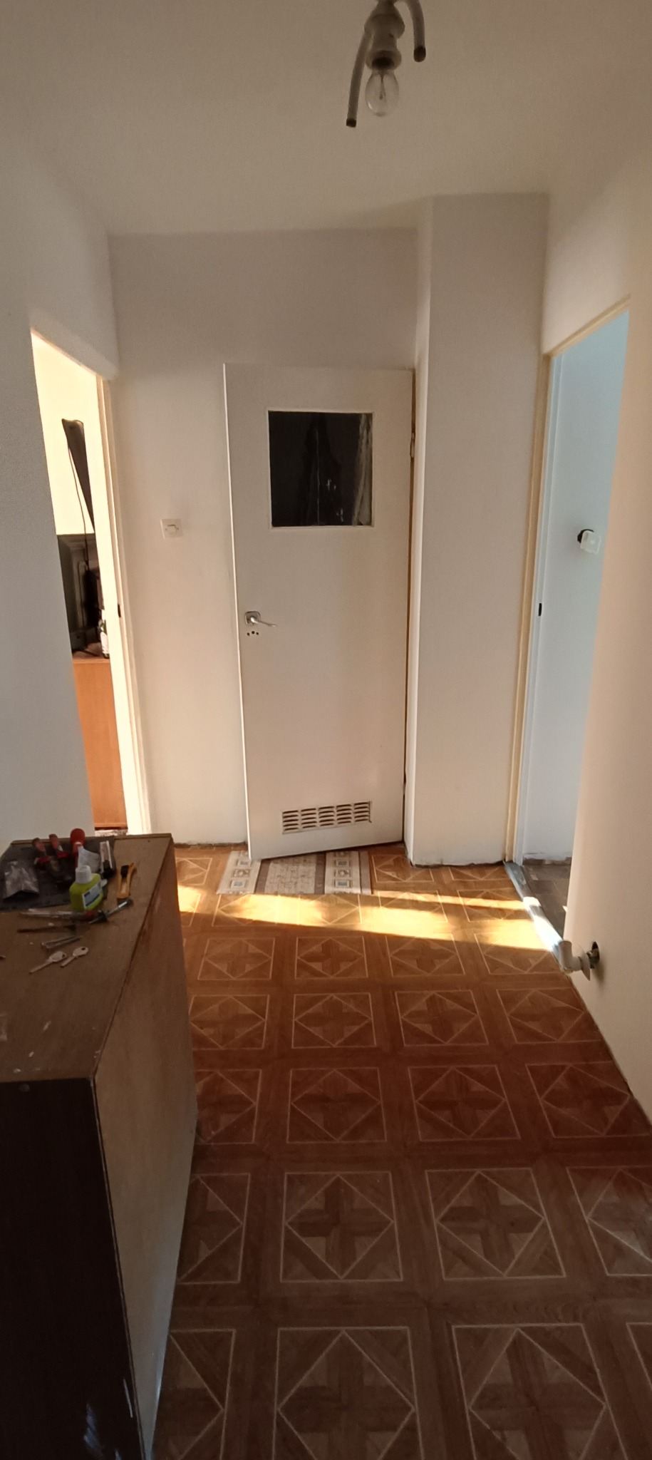 Korytarz w trakcie odnawiania z widocznymi narzędziami, białą ścianą, drzwiami z szybką i wzorzystą podłogą z płytek.