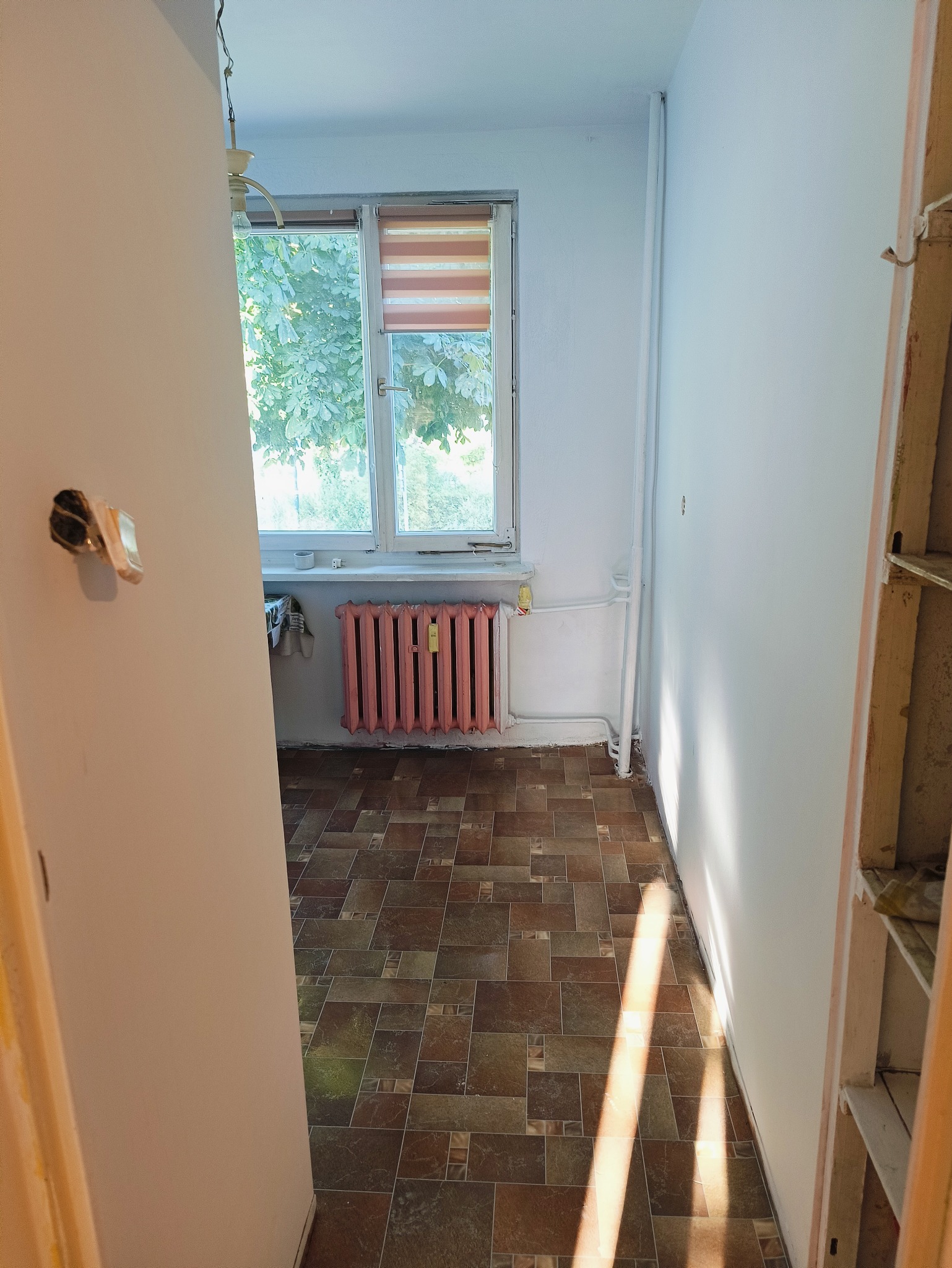 Pokój w trakcie odświeżania, widoczne nowe płytki podłogowe, pomalowany na różowo kaloryfer i świeżo pomalowane ściany na biało, przez okno widać zieleń drzew.