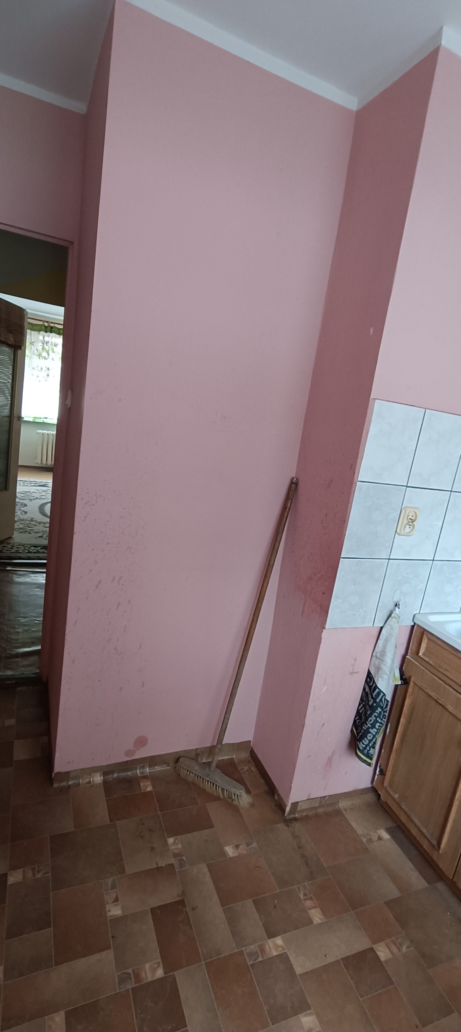 Różowa ściana w trakcie przygotowań do malowania, widoczne ślady zabrudzeń i szpachli, oparta o ścianę miotła, fragment płytek i szafki kuchennej w tle.
