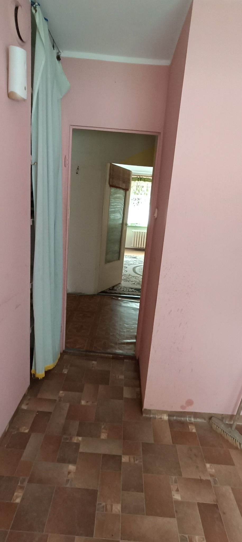 Widok z korytarza na pokój z widoczną podłogą w brązowych odcieniach, różowe ściany i drzwi z zasłoną.