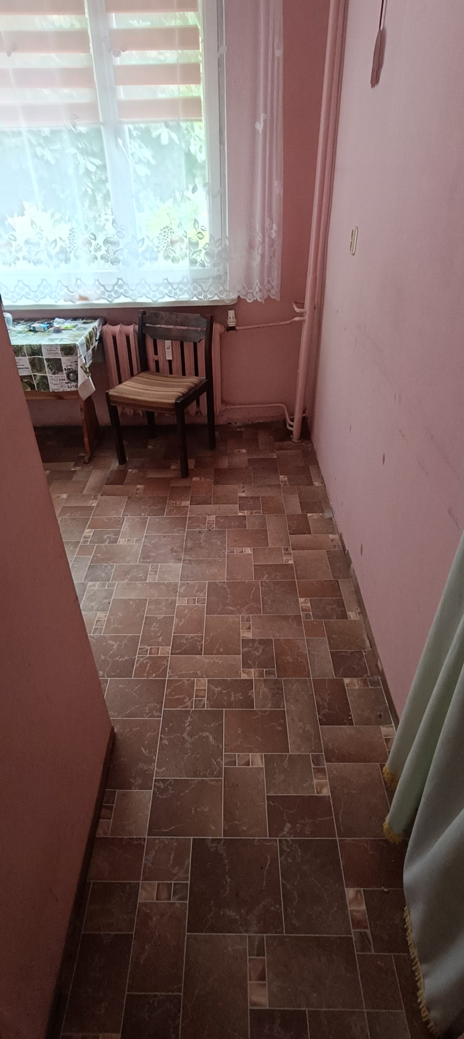 Pokój z brązową podłogą z płytek imitujących kamień, różowe ściany, okno z białymi firankami i drewniane krzesło. Widoczne rury grzewcze przy ścianie.