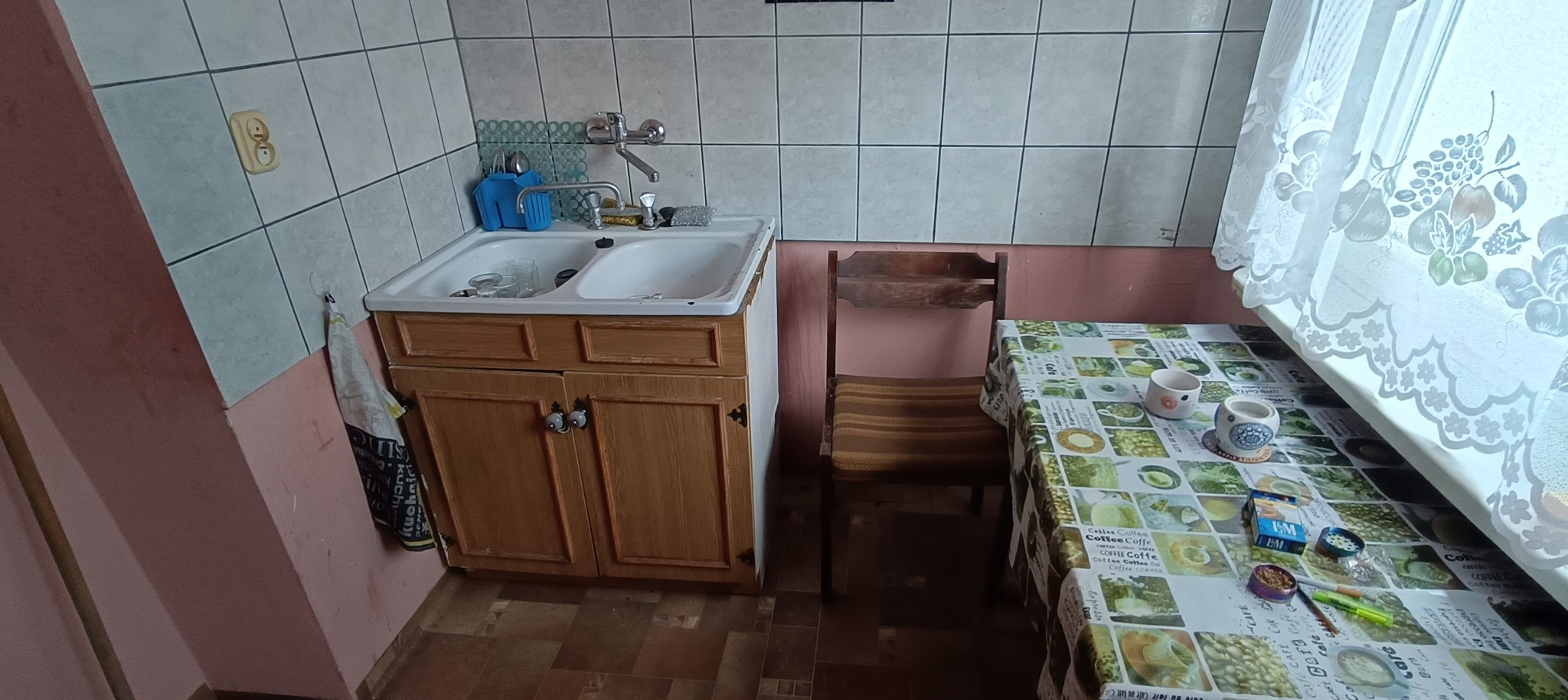 Umywalka z szafką w kuchni przed remontem, widoczne stare płytki, drewniane szafki, krzesło i stół nakryty ceratą z motywem kawy.