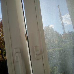 SPRZĄTANIE Z JUSTYNĄ JUSTYNA KRÓLAK - Otwarta biała rama okienna z widocznym drzewem z jesiennymi liśćmi po lewej stronie i brudną szybą z widocznym kominem przemysłowym po prawej.