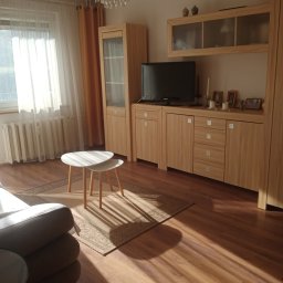 SPRZĄTANIE Z JUSTYNĄ JUSTYNA KRÓLAK - Jasne wnętrze salonu z drewnianą podłogą, meblami z jasnego drewna, białymi stolikami kawowymi i dywanem w neutralnych barwach, oświetlone naturalnym światłem wpadającym przez okno z białymi...