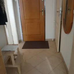 Przedpokój w Kętrzynie z drewnianymi drzwiami wejściowymi, brązowym dywanikiem i beżową podłogą z płytek. Widoczna drabinka z dokumentami i fragment lustra w drewnianej ramie.