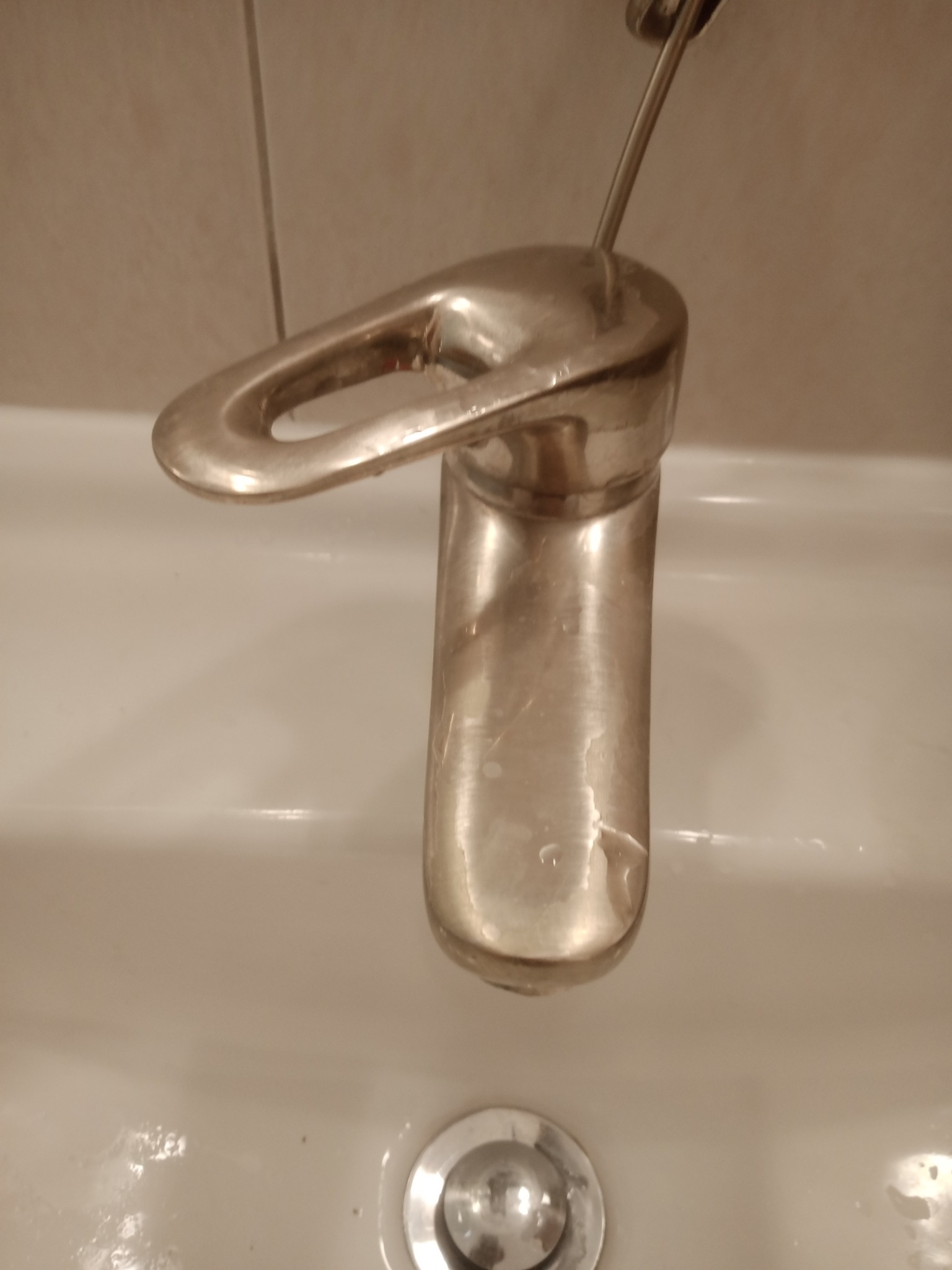 Błyszcząca, chromowana bateria umywalkowa z widocznymi zaciekami na korpusie, osadzona w białej umywalce, na tle beżowych płytek.