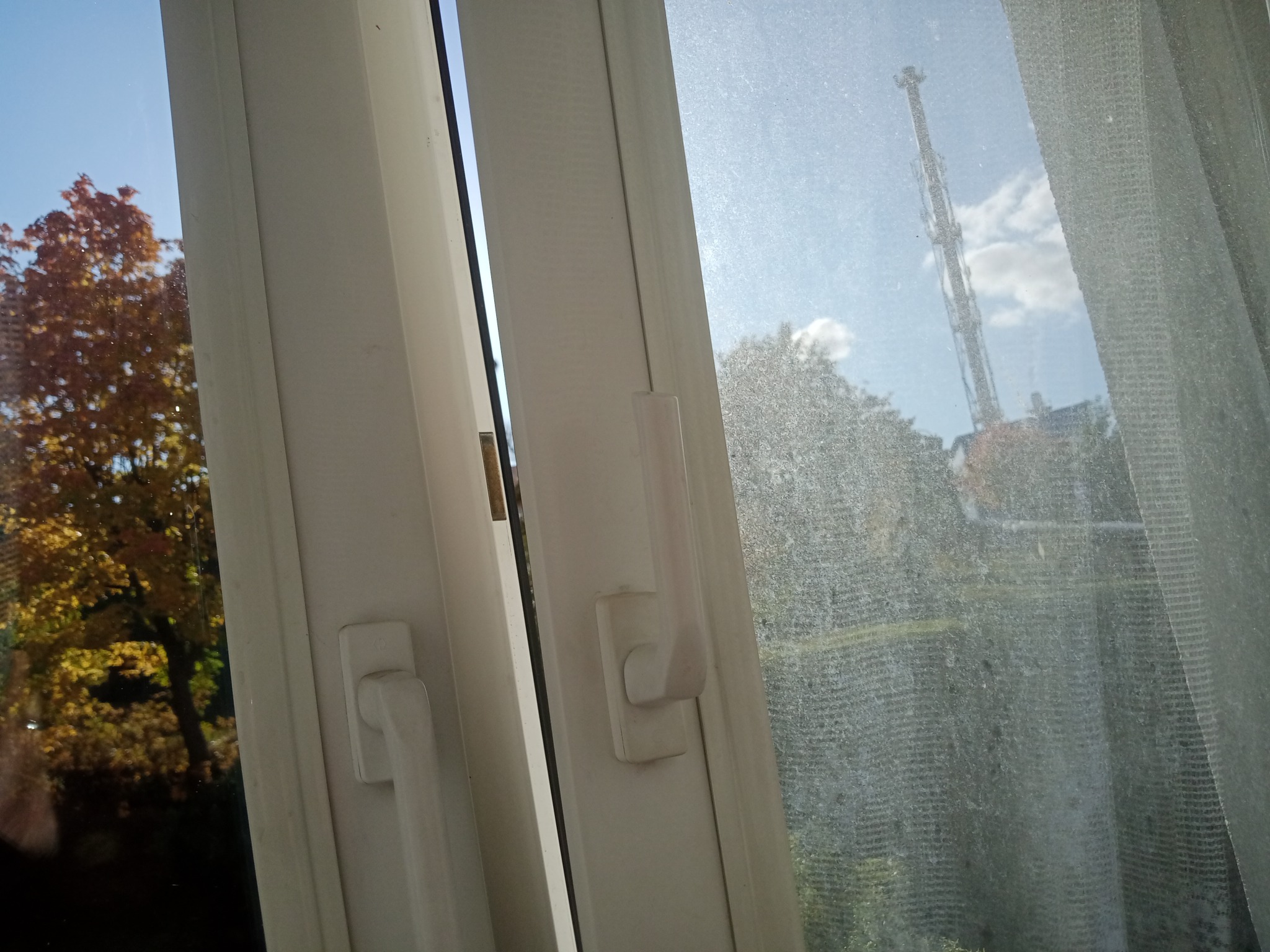 Otwarta biała rama okienna z widocznym drzewem z jesiennymi liśćmi po lewej stronie i brudną szybą z widocznym kominem przemysłowym po prawej.