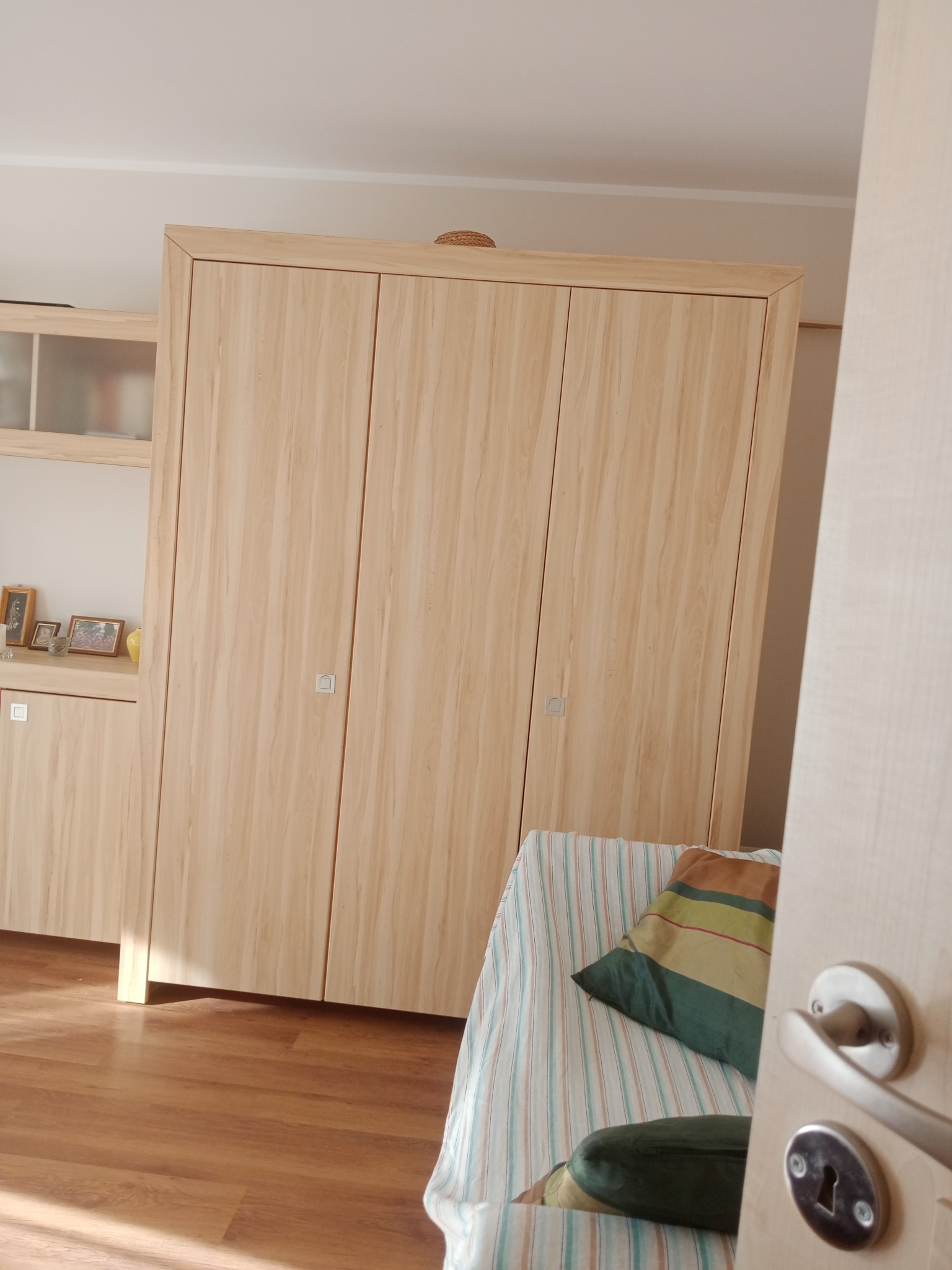 Jasne, drewniane, trzydrzwiowe szafy wnękowe z kwadratowymi uchwytami, widoczne z perspektywy otwartych drzwi, obok łóżka z poduszkami.