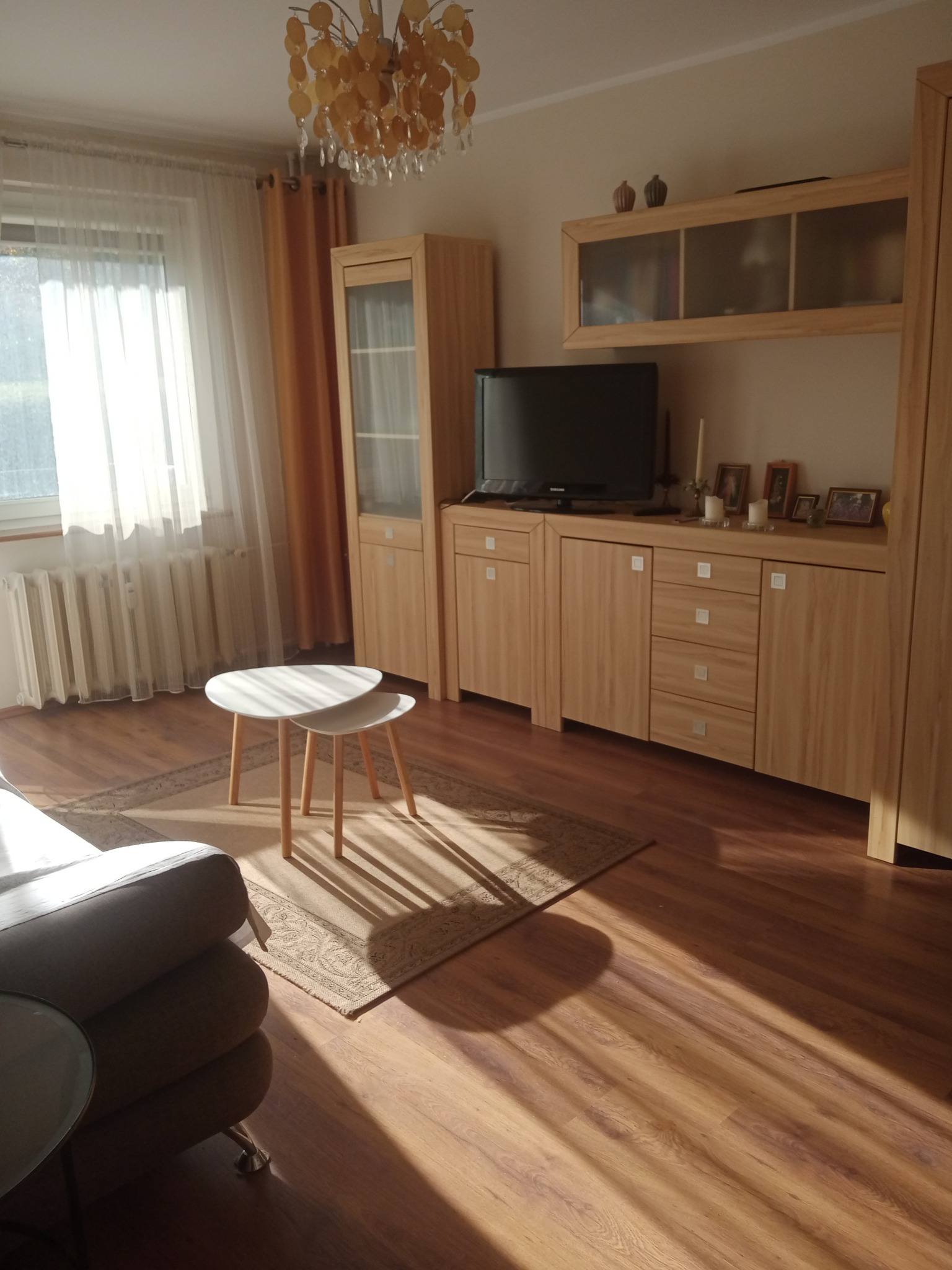 Jasne wnętrze salonu z drewnianą podłogą, meblami z jasnego drewna, białymi stolikami kawowymi i dywanem w neutralnych barwach, oświetlone naturalnym światłem wpadającym przez okno z białymi...