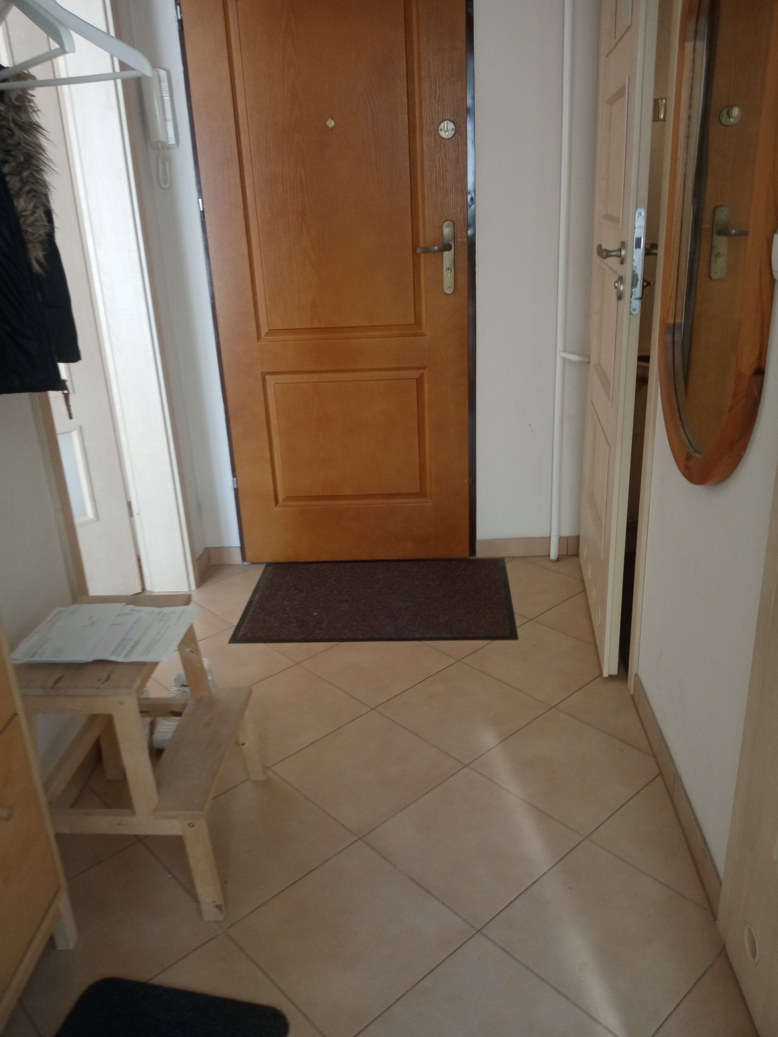Przedpokój w Kętrzynie z drewnianymi drzwiami wejściowymi, brązowym dywanikiem i beżową podłogą z płytek. Widoczna drabinka z dokumentami i fragment lustra w drewnianej ramie.