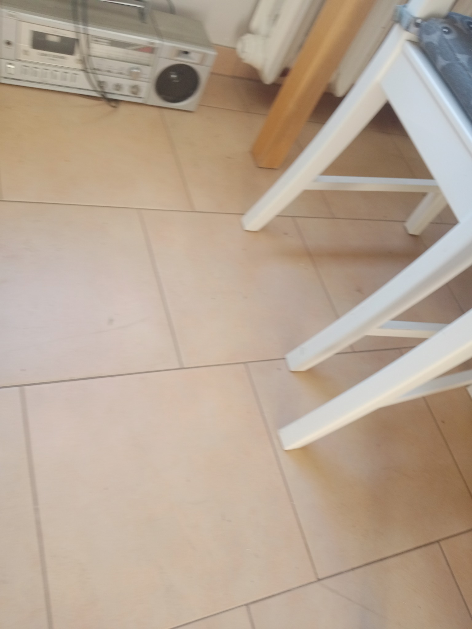 Beżowe płytki podłogowe z widocznymi nogami białego krzesła i fragmentem starego, srebrnego boomboxa w tle.