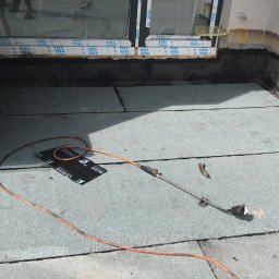 Gacki Dach - Montaż papy termozgrzewalnej na dachu płaskim, widoczny palnik dekarski, fragment okna z okleiną, oraz resztki materiałów izolacyjnych przy krawędzi dachu.