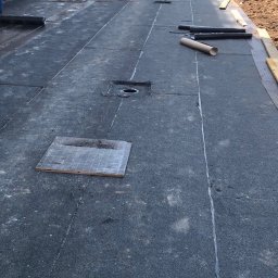 Gacki Dach - Widok z góry na płaski dach pokryty czarną papą termozgrzewalną z widocznymi miejscami zgrzewania, kominkami wentylacyjnymi i narzędziami pracy, na tle budynków w stanie surowym.