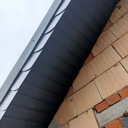 Gacki Dach - Fragment dachu z blachy trapezowej w kolorze grafitowym, widoczny od dołu, przylegający do muru z cegły dziurawki i betonu, wykonany pod kątem, na tle pochmurnego nieba.