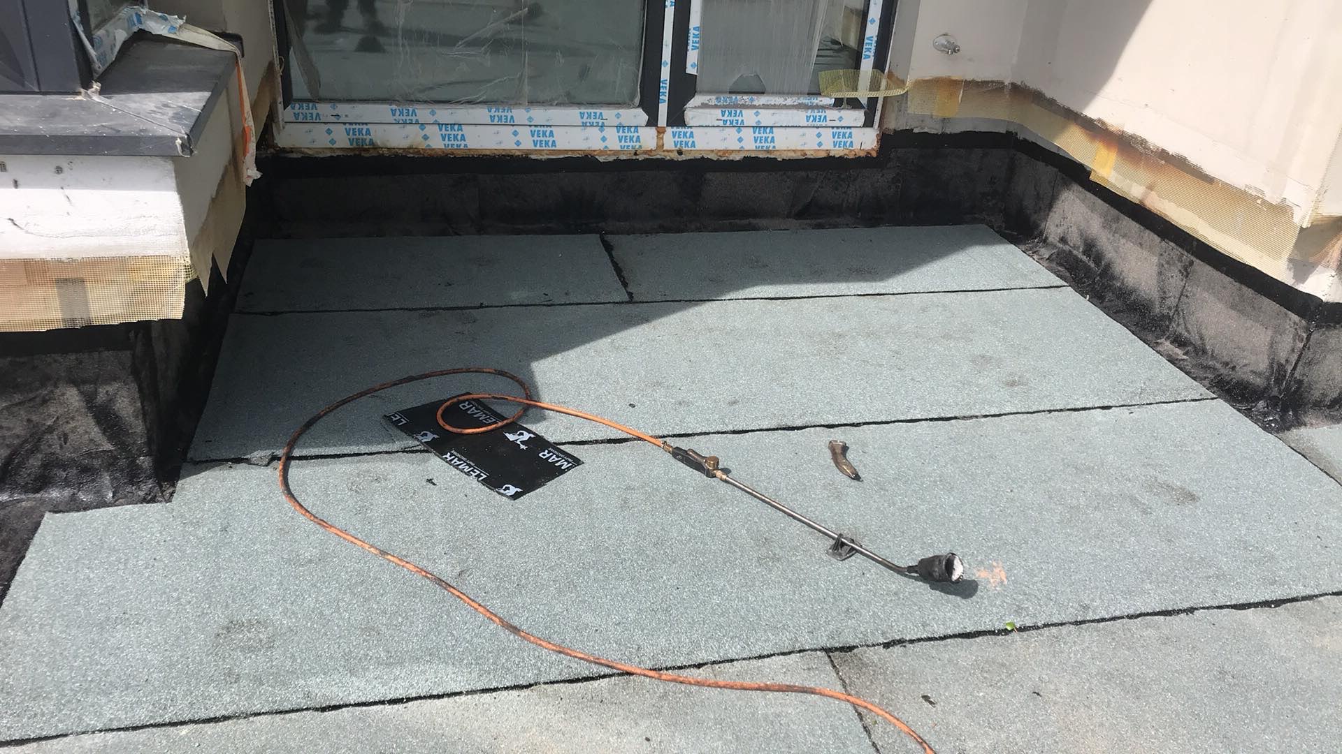 Montaż papy termozgrzewalnej na dachu płaskim, widoczny palnik dekarski, fragment okna z okleiną, oraz resztki materiałów izolacyjnych przy krawędzi dachu.