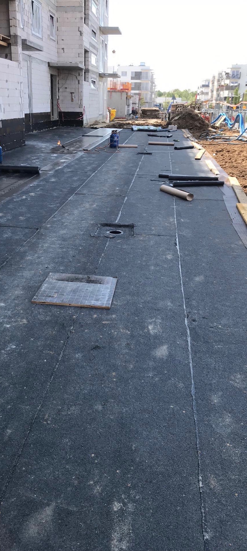 Widok z góry na płaski dach pokryty czarną papą termozgrzewalną z widocznymi miejscami zgrzewania, kominkami wentylacyjnymi i narzędziami pracy, na tle budynków w stanie surowym.