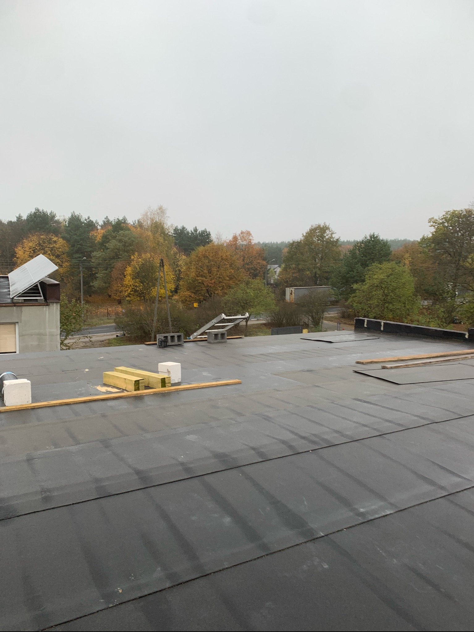 Płaski dach pokryty papą termozgrzewalną w trakcie prac, widoczne deski, bloczki betonowe, drabina w tle, jesienny krajobraz