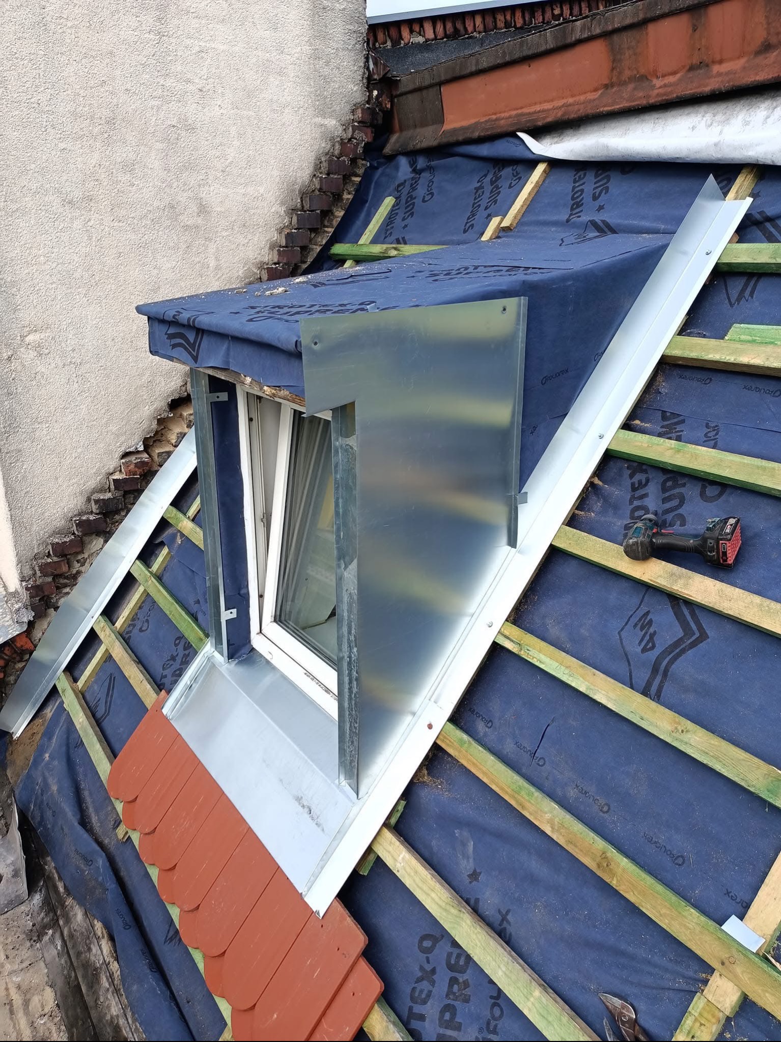 Montaż okna dachowego z obróbką blacharską, widoczne elementy konstrukcji dachu, membrana dachowa i częściowo ułożona dachówka ceramiczna, w tle ściana budynku.