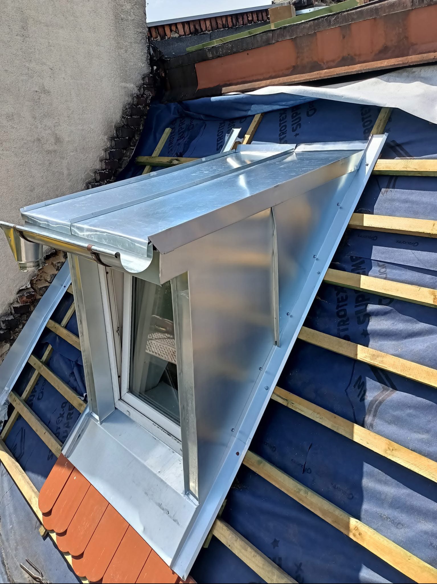 Nowa, metalowa obróbka blacharska okna dachowego, z widoczną rynną, na tle warstwy hydroizolacyjnej i drewnianej konstrukcji dachu, z fragmentem starego pokrycia dachowego.