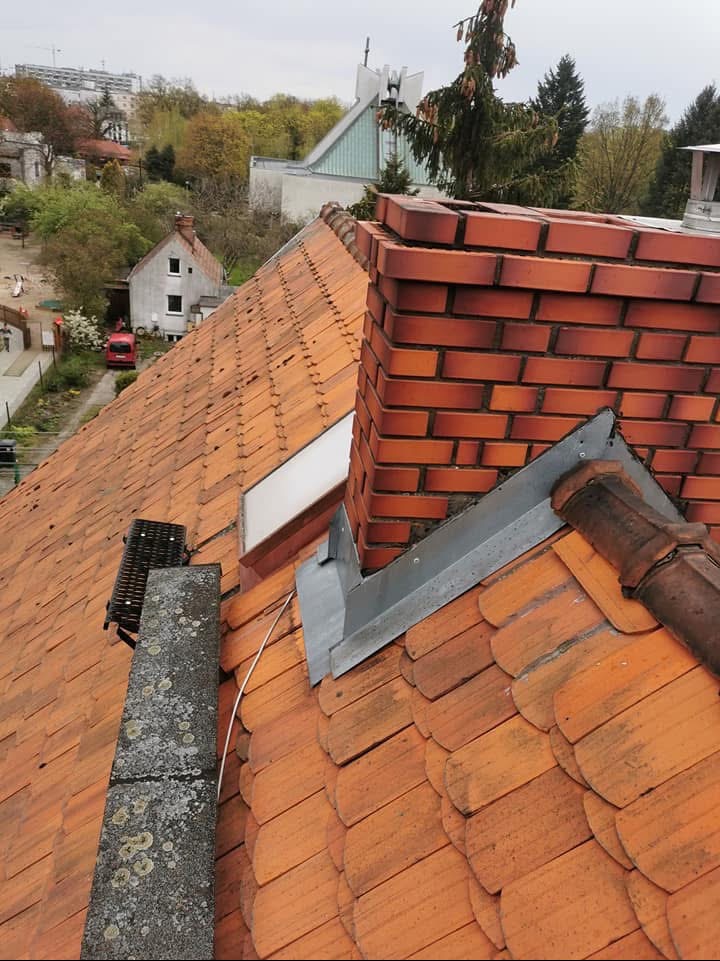 Ceglany komin z obróbką blacharską na dachu z dachówki karpiówki z widocznym oknem dachowym i elementami systemu odśnieżania, w tle zabudowa miejska i drzewa.