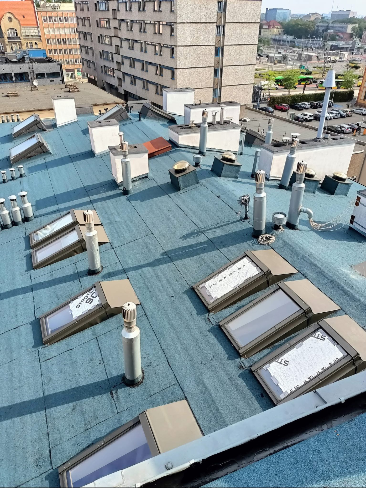 Widok z góry na płaski dach pokryty papą w kolorze niebieskim z zamontowanymi świetlikami dachowymi, kominami wentylacyjnymi i obróbkami blacharskimi, w tle zabudowa miejska.