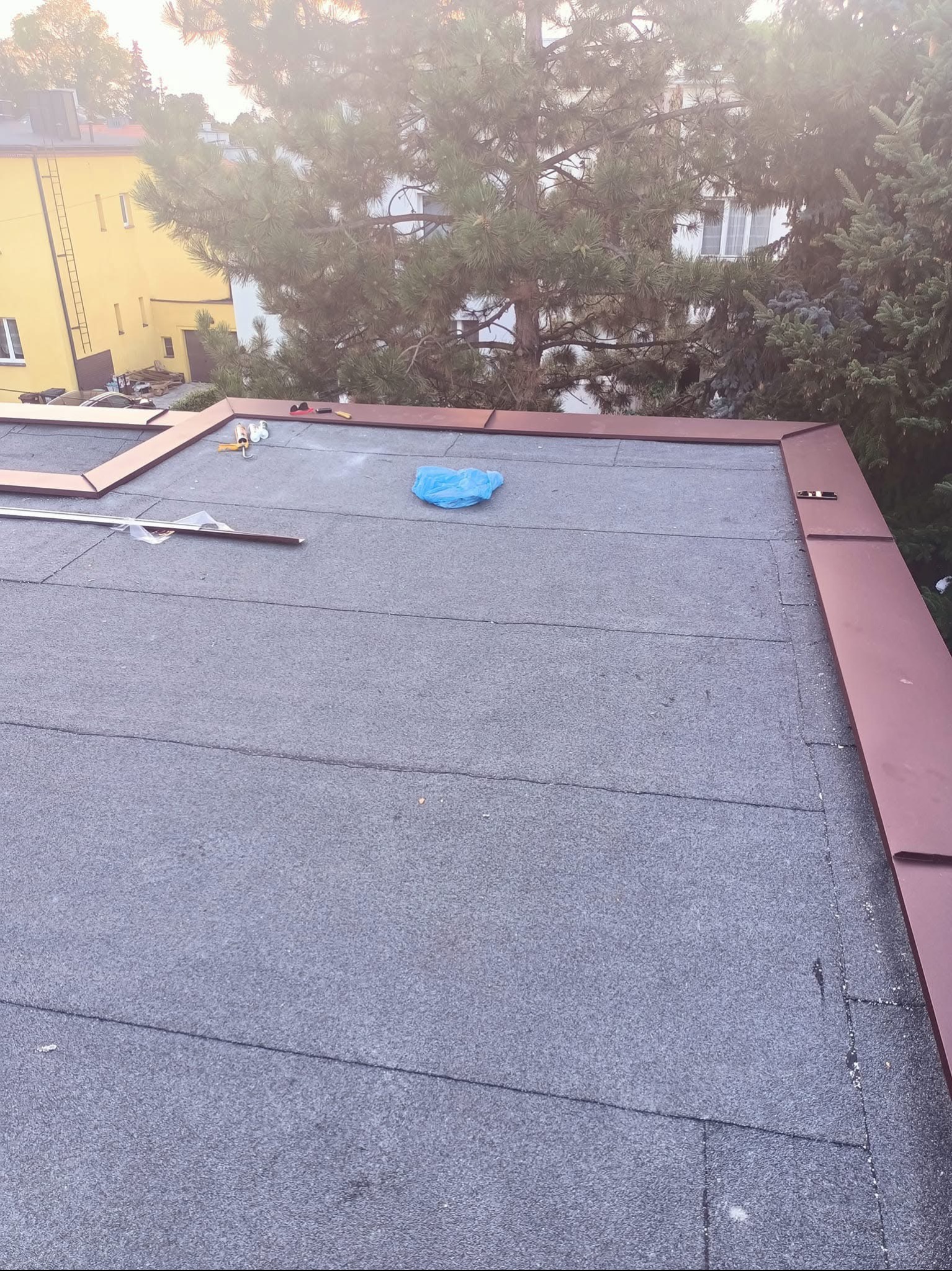 Widok z góry na płaski dach pokryty papą termozgrzewalną z przygotowanymi elementami obróbek blacharskich w kolorze brązowym i narzędziami, na tle drzew i budynku.