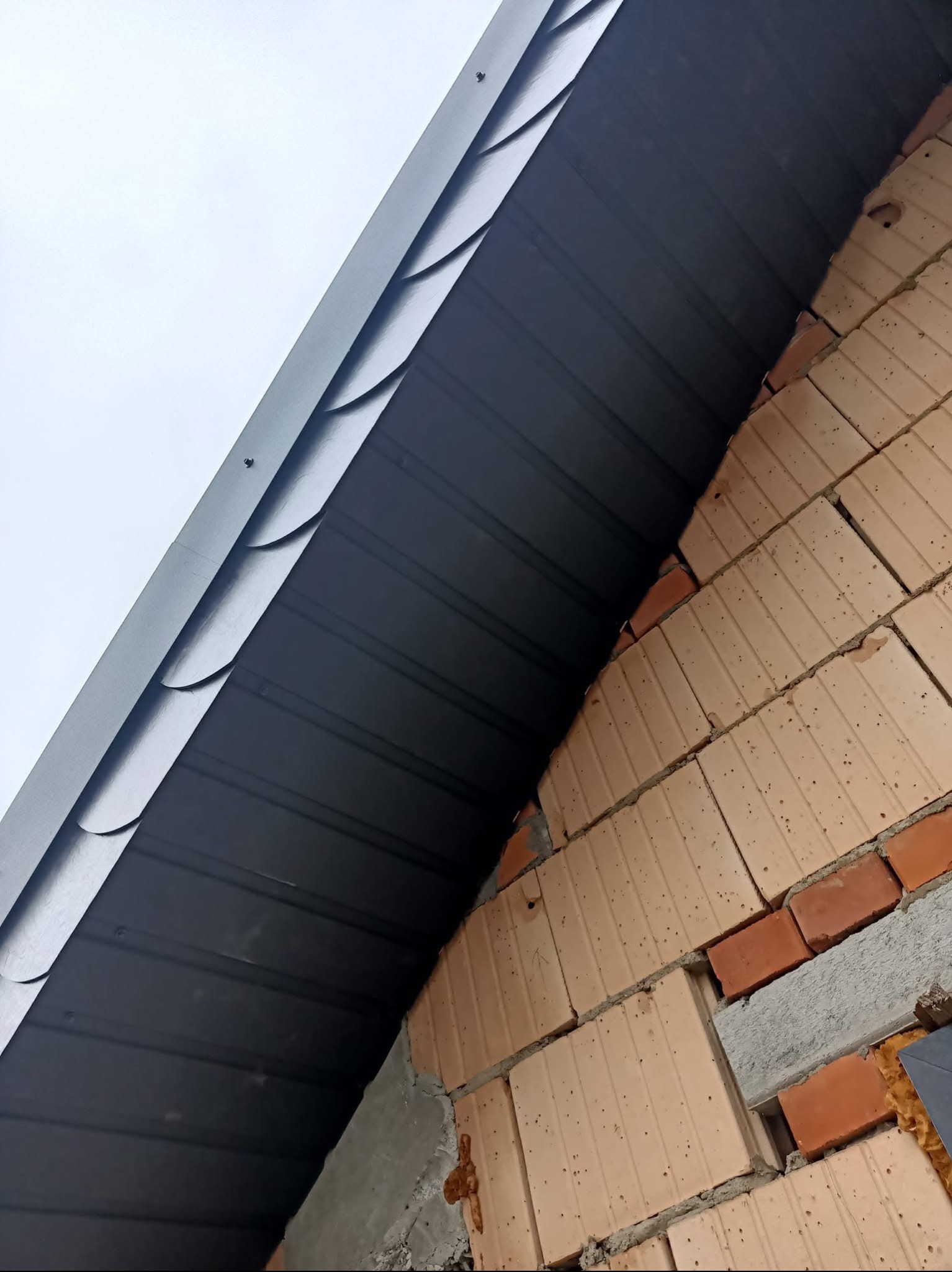 Fragment dachu z blachy trapezowej w kolorze grafitowym, widoczny od dołu, przylegający do muru z cegły dziurawki i betonu, wykonany pod kątem, na tle pochmurnego nieba.