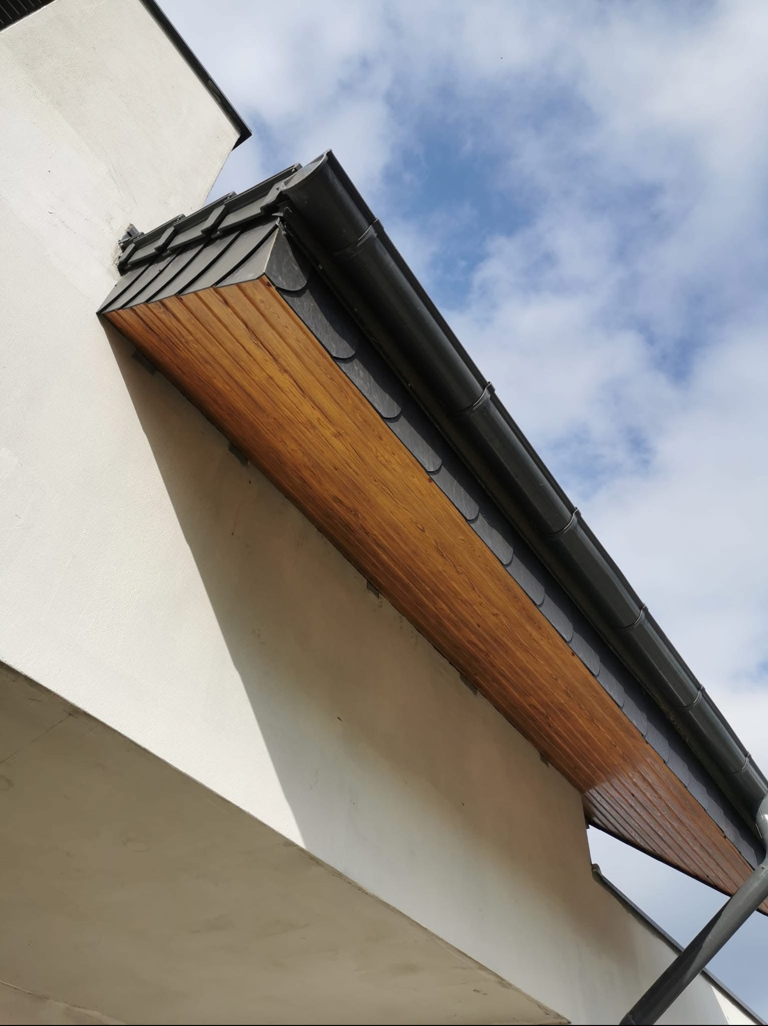 Zamontowana, szara rynna z drewnianą podbitką dachową i fragmentem dachu z dachówki bitumicznej, widoczne na tle błękitnego nieba z chmurami.