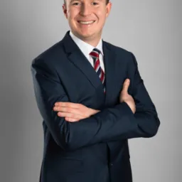 Uśmiechnięty mężczyzna w granatowym garniturze, białej koszuli i krawacie w paski, stoi na szarym tle z założonymi rękami.