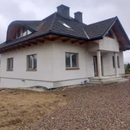 Nowoczesny dom jednorodzinny z ciemną dachówką i lukarnami o nietypowym, zaokrąglonym kształcie. Elewacja w jasnym odcieniu. Widoczny fragment terenu przed budynkiem.