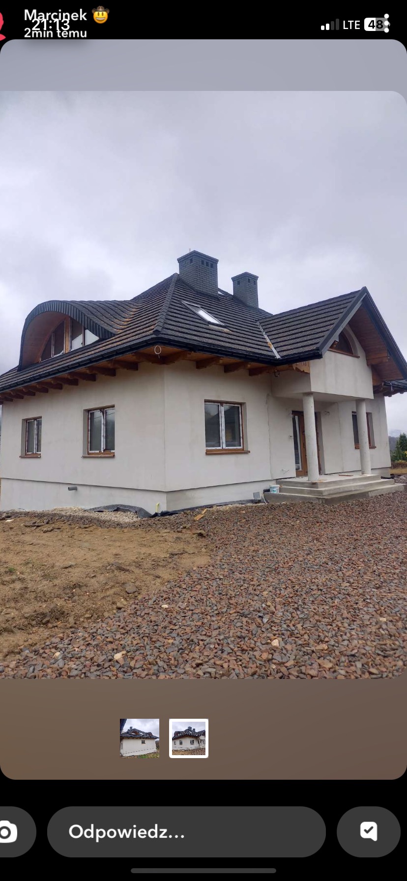 Nowoczesny dom jednorodzinny z ciemną dachówką i lukarnami o nietypowym, zaokrąglonym kształcie. Elewacja w jasnym odcieniu. Widoczny fragment terenu przed budynkiem.