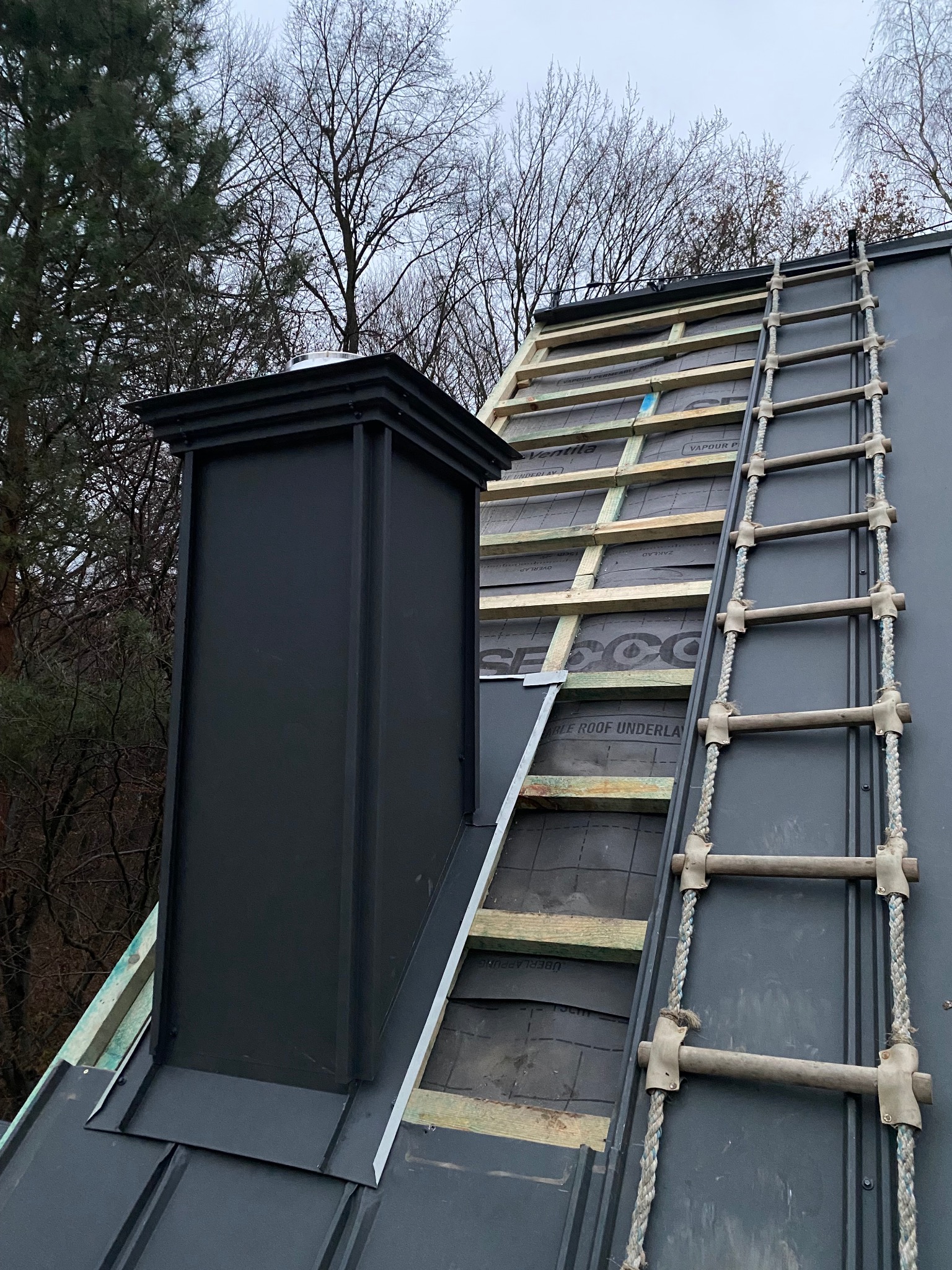 Czarny komin z blachy na dachu z drabinką dekarską. Widoczna warstwa izolacyjna i drewniana konstrukcja dachu w trakcie prac. Tło stanowią drzewa.