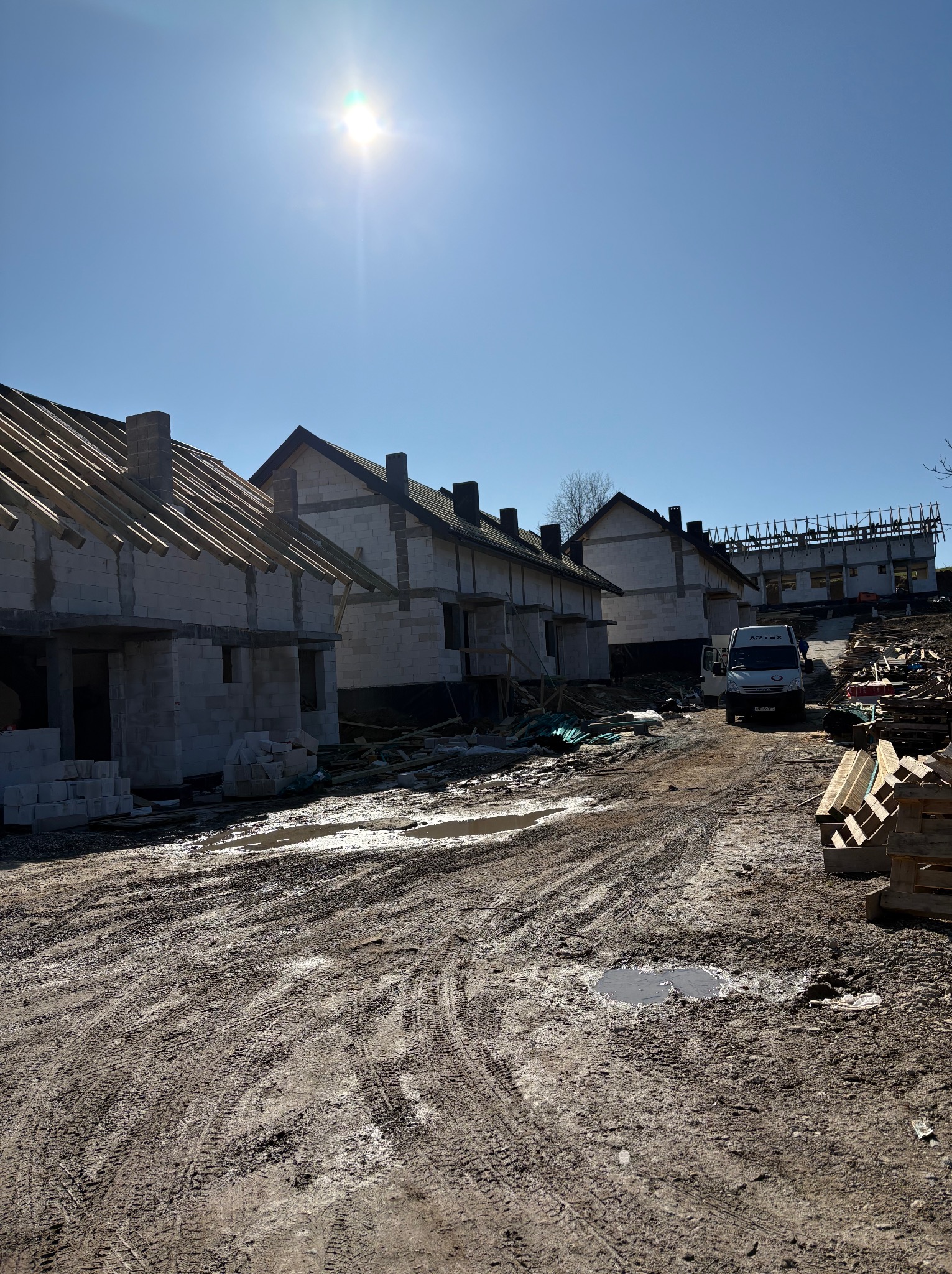Surowe domy w zabudowie szeregowej z częściowo wykonaną więźbą dachową, na błotnistym terenie budowy w słoneczny dzień. Widoczny samochód dostawczy.