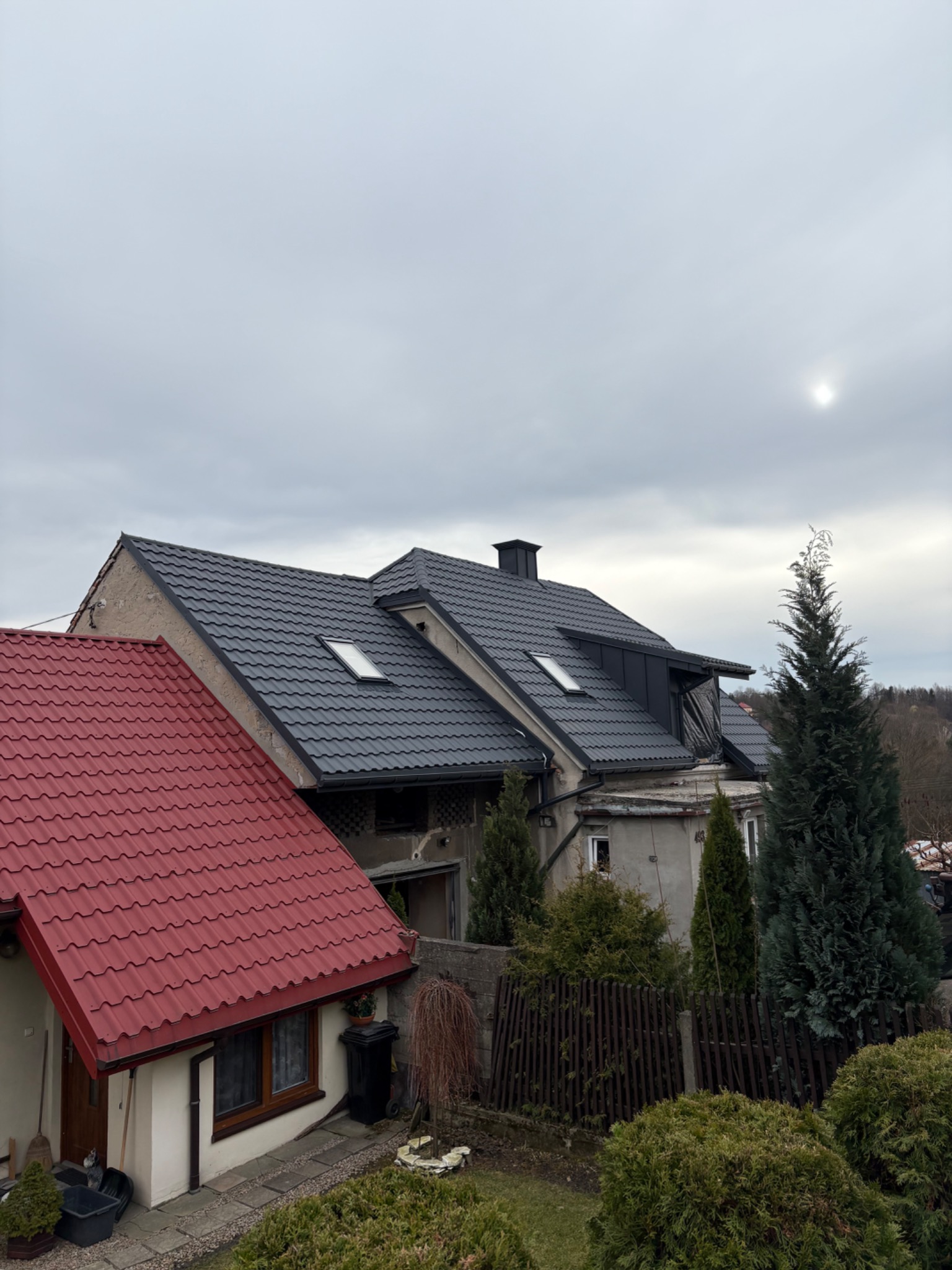 Dom z ciemną dachówką i oknami dachowymi w Mszanie Dolnej. Na pierwszym planie fragment dachu z czerwoną dachówką. Pochmurne niebo w tle.